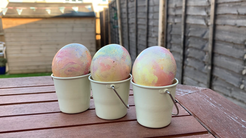 Marmorerte egg med hint av pastellfarger som grønn, blå, rosa og gul som ligger i hver sin lille, hvite bøtte/eggholder.
